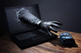 EIne Hand im schwarzen Handschuh greift aus einem Laptop die auf dem Tisch befindliche Geldbörse.