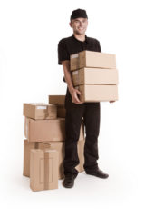 Mann in schwarzer Kleidung steht vor vielen Paketen und hat selbst drei Pakete in der Hand.