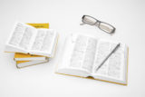 Aufgeschlagenes Langenscheidt Wörterbuch auf dem ein Kugelschreiber liegt. Darüber liegt eine Brille und links daneben befinden sich weitere aufeinander gestapelte Langenscheidt Wörterbüchern, wobei das oberste ebenfalls aufgeschlagen ist.