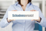 Frau in blauer Bluse hebt ein weißes Schild mit der Aufschrift "Schadensersatz" in den Händen.