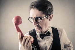 Ein Mann mit Brille hat einen roten Telefonhörer in der Hand und schaut diesen genervt an.