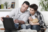 Vater und Sohn sitzen vor einem Laptop und essen Popcorn.