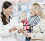 Apothekerin übergibt Medikament an Frau mit Kind auf dem Arm