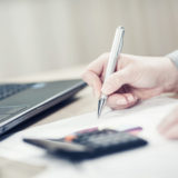 Rechte Hand die mit einem Kugelschreiber Notizen macht während auf dem Blatt ein taschenrechner liegt, und im Hintergrubnd ein Laptop zu sehen ist