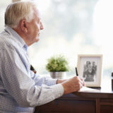 Älterer Mann, der am Schreibtisch sitzt und schreibt, im Hintergrund Familienfoto