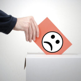 Person wirft eine rote Karte mit einem traurigen Smilie in eine Wahlbox
