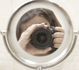 Frau mit Fotokamera schießt Foto, ihr gegenüber steht ein Spiegel