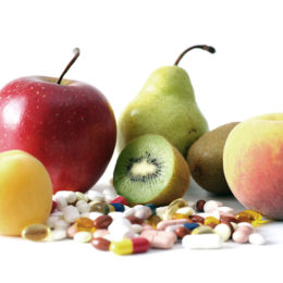 viele bunte Tabletten, die vor einem Apfel, einer Birne, einer mittig durchgeschnittenen Kiwi, und einem Pfirsich liegen
