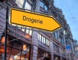 Gelbes Straßenschild mit der Aufschrift "Drogerie" welches nach rechts weist.