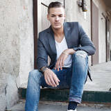 Mann, gekleidet in Jeans und Jacket, sitzt auf einer Treppenstufe und post. Er trägt eine Uhr an seinem rechten Arm und ein Bildernes Armband an seinem linken Handgelenk