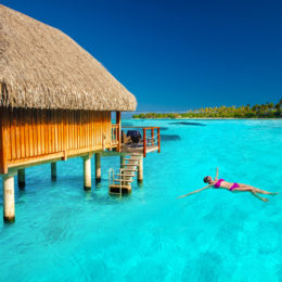 Eine Frau lässt sich in einem pinken Bikini im türkisen Meer treiben. Neben ihr befindet sich ein Bungalow mit Zugang zum Wasser. Im Hintergrund ist das Ufer und strahlend blauer Himmel zu sehen.