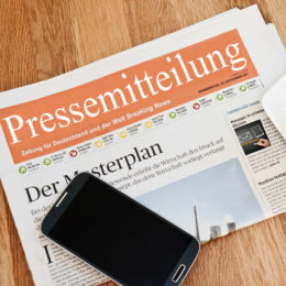 Zeitung, auf der der in weiß gehaltene Schriftzug 'Pressemitteilung' auf orangenem Hintergrund besonders hervorsticht. Auf der Zeitung liegt ein schwarzes Smartphone