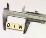 3 Buchstabenwürfel die das Wort "DIN" zeigen, eingeklemmt in einem Lineal