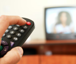 Hand, die eine Fernbedienung hält, im Hintergrund ein Fernseher