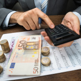 Geschäftsmann mit Taschenrechner und Kugelschreiber in der Hand an einem Tisch, auf dem Geldscheine, Münzen und eine Tabelle liegen