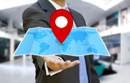 Mann im Anzug hebt eine blaue Landkarte in der Hand, auf die ein roter Standortpunkt zeigt.