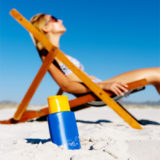 Sonnenmilch in blauem Design steckt vor einer im Liegestuhl liegenden Frau im Sand.