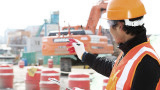 Bauarbeiter in oranger Kleidung hat ein Klemmbrett in der Hand und zeigt auf eine im Hindergrund befindliche Baustelle.