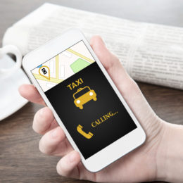 Hand hält ein Smartphone mit dem es ein Taxi ruft. Im Hintergrund befindet sich eine Zeitung und eine Kaffeetasse
