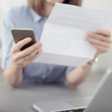 Eine Frau sitzt an einem weißen Tisch. Sie hält in der linken Hand einen Brief und in der rechten Hand ein Smartphone