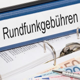 Ordner mit Aufschrift "Eundfunkgebühren" und Geldscheinen im Vodergrund