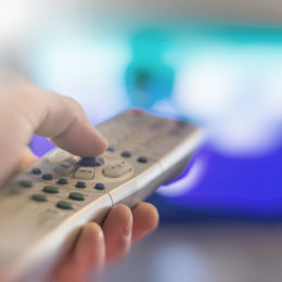 Eine Hand hält eine Fernbedienung, im Hintergrund läuft ein Fernseher