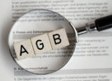 Drei Würfel mit der Aufschrift "AGB" liegen unter einer Lupe auf Papier.