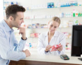ein männlicher Kunde wird von einer jungen Apothekerin beraten, im Hintergrund sieht man Regale mit Medikamenten