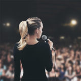 Frau steht mit einem Mikrofon in der rechten Hand vor einem großen Publikum auf der Bühne