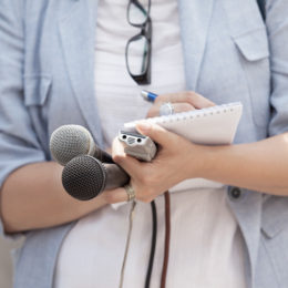 eine Journalistin hält Mikrofone und ein Diktiergerät in der Hand, während sie sich Notien auf einem Block macht