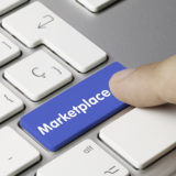 Der Zeigefinger einer Person drückt auf eine blau hinterlegte Taste mit der Aufschrift "Marketplace" auf einer silbernen Tastatur mit sonst weißen Tasten