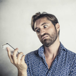 Mann mit ausgefallenem Hemd vor weiß-grauem Hintergrund schaut gelangweilt auf sein in der Hand liegendes Smartphone