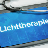 Der Schriftzug "Lichttherapie" erscheint in weißen Buchstaben auf dem Display eines Tablets. Daneben liegt ein Stethoskop.