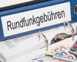 blauer Ordner mit der Aufschrift "Rundfunkgebühren" auf einem augeklappten Ordner mit diversen Dokumenten und Geldscheinen