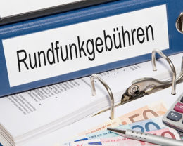 blauer Ordner mit der Aufschrift "Rundfunkgebühren" auf einem augeklappten Ordner mit diversen Dokumenten und Geldscheinen