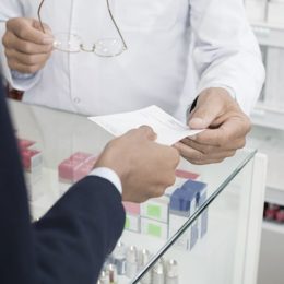 Apotheker übergibt einem Kunden über den Tresen hinweg ein beschriftetes Stück und hält in der anderen Hand eine Brille
