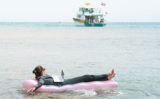 junge Frau liegt mit ihrem Laptop auf einer Luftmatratze im Meer, im hintergrund eine Yacht