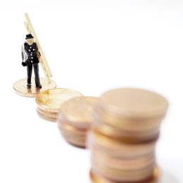 Figur eines Miniatur-Schornsteinfegers, der auf einem Stapel von Geldmünzen steht und noch weitere vor sich hat