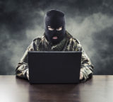 maskierter Mann, der symbolisch für einen Internetverbrecher steht, sitzt vor dunklem, verrauchten Hintergrund am Laptop