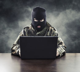 maskierter Mann, der symbolisch für einen Internetverbrecher steht, sitzt vor dunklem, verrauchten Hintergrund am Laptop