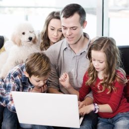 Eine vierköpfige Familie - Vater, Mutter, Sohn, Tochter - sitzt auf einem Sofa vor dem Laptop