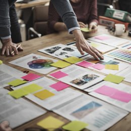 Geschäftsmänner beugen sich über einen Tisch, auf dem sie mit Post-Its versehene Unterlagen ausgebreitet haben, brainstorming