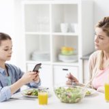 Mutter sitzt mit ihrem Kind beim Essen; die Tochter starrt auf ihr Smartphone, die Mutter schaut traurig