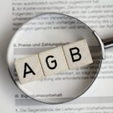 Drei Würfel mit den Buchstaben "AGB" liegen auf einem Vertragswerk; darüber wird eine Lupe gehalten