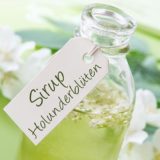 ein aus der Holunderblüte erzeugter Sirup in weißer Glasflasche mit angefügtem Label "Sirup Holunderblüten"; im Hintergrund: eine Holunderblüte