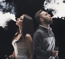 Mann und Frau stehen Rücken an Rücken während beide eine gewaltige Dampfwolke ausatmen, die durch einen Vaporizer erzeugt wurde