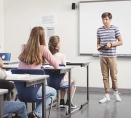 Junge steht vor einer Schulklasse und hält ein Referat. Hinter ihm ist ein Whiteboard.
