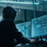 Mann mit dunklem Kapuzenpullover und hochgezogener Kapuze sitzt in einem schwach beleuchteten Raum vor einem Computer-Bildschirm