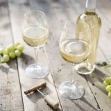 Zwei Weingläser mit Weißwein gefüllt und eine Weinflasche auf Holz-Boden. daneben Trauben und ein Korkenzieher