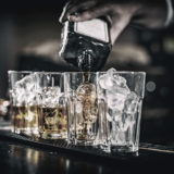 Barkeeper schenkt Whiskey in vier Gläser mit Eiswürfel